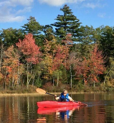 Woman in a red kayak with blue life jacket on a lake in fall.