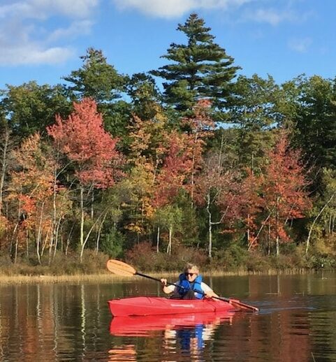 Woman in a red kayak with blue life jacket on a lake in fall.