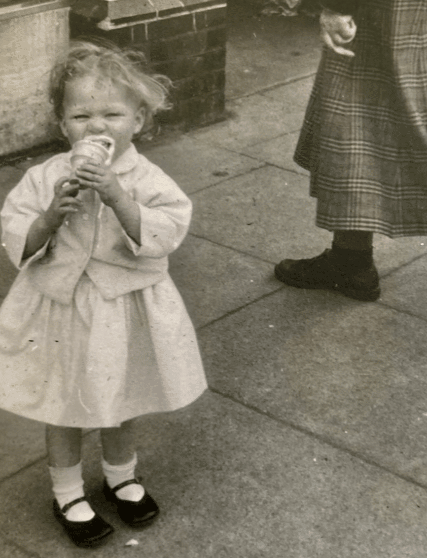 Little girl, aged two, in a dress, sweater, and Mary-Janes and socks, eating a messy ice cream cone.