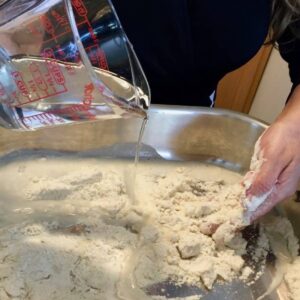 Close-up of a woman's hands adding water to flour to make pupusa dough in a silver mixing bowl.
