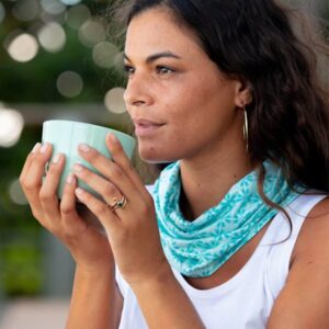A woman wearing a turquoise Ripskirt scarf around her neck, holding a cup and gazing in the distance.