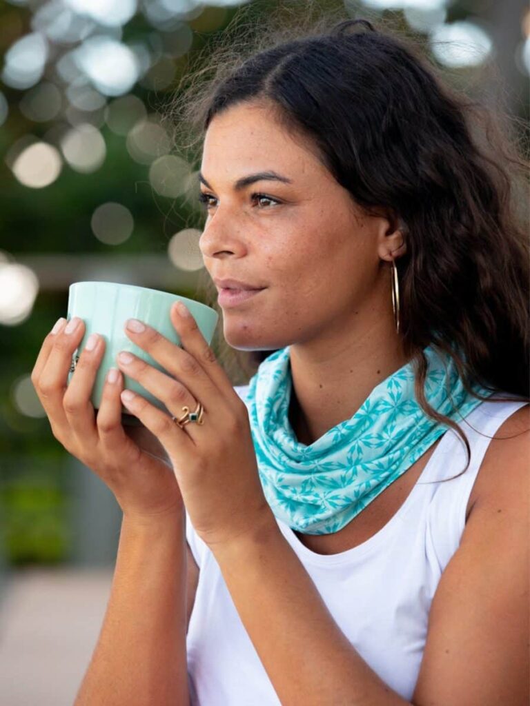 A woman wearing a turquoise Ripskirt scarf around her neck, holding a cup and gazing in the distance.