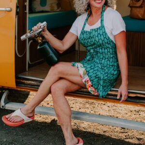 Smiling woman wearing a green apron holding a culinary torch outside an RV.