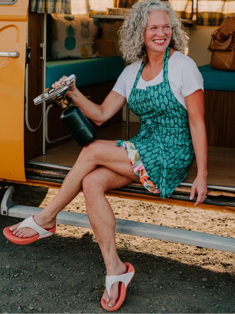 Smiling woman wearing a green apron holding a culinary torch outside an RV.